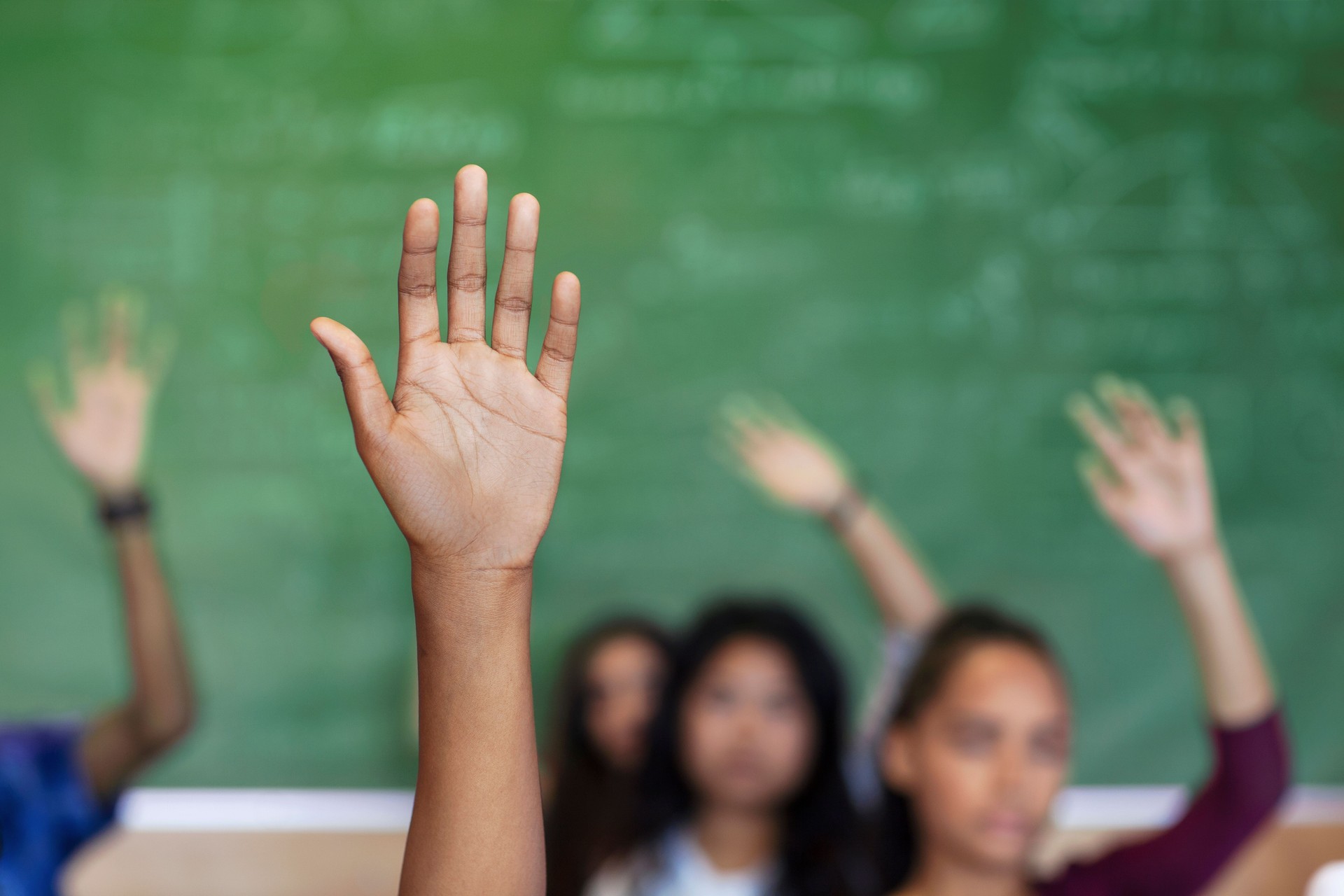 Diverse multi-cultural teenagers with hands raised sitting in classroom